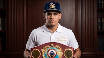 Un hombre con gorra azul del WBC y camisa polo blanca sostiene un cinturón de boxeo de la WBO frente a un mueble de madera.