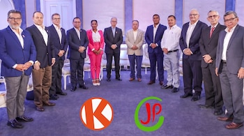 Once personas, diez hombres y una mujer, visten ropa formal y posan de pie en un salón. En el suelo se ven los logos K naranja y JP rojo y verde.