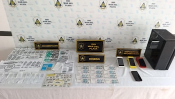 Una mesa blanca con signos de policía, documentación, recibos, billetes de dinero, teléfonos celulares y una computadora de escritorio