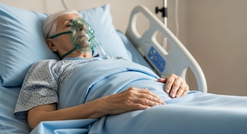Un adulto mayor recostado en una cama de hospital con bata y manta azules. Tiene una mascarilla de oxígeno verde. Sus manos están sobre la manta.