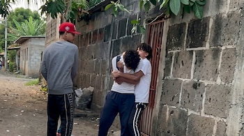 Tres jóvenes en un camino de tierra junto a una pared de bloques; dos se abrazan mientras el tercero observa. Hay vegetación tropical al fondo.