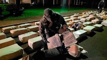 Un agente de seguridad con gorra y guantes examina paquetes envueltos en cinta rodeado de decenas de bultos rectangulares en la calle de noche