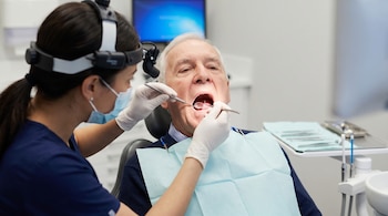 Una dentista con gorro de lupa y mascarilla examina la boca abierta de un hombre mayor sentado en un sillón dental, utilizando un espejo y una sonda.
