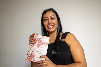 Mujer con cabello oscuro y labios rojos sonríe a la cámara mientras sostiene un libro blanco con diseño de flores rojas y el texto '9 hábitos japoneses'