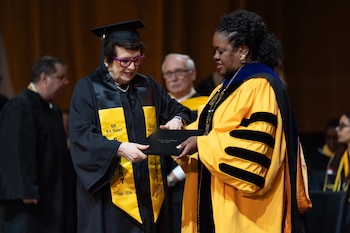 Dos mujeres en togas académicas en un escenario. La de la izquierda, con birrete negro, recibe un diploma de la de la derecha, con toga amarilla y azul