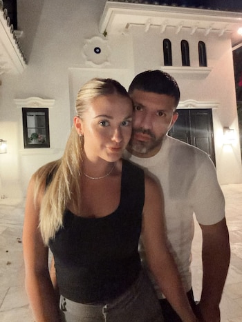 Una mujer y un hombre posan de pie frente a un edificio blanco iluminado por la noche. La mujer, de pelo rubio recogido, usa camiseta negra. El hombre usa camiseta blanca