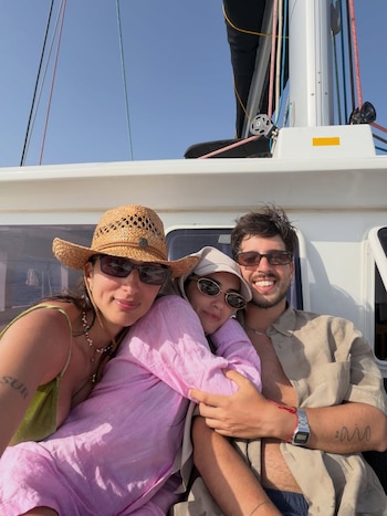 Tres personas jóvenes con gafas de sol, dos mujeres y un hombre, sonríen a cámara mientras están sentadas en la cubierta de un velero bajo el sol