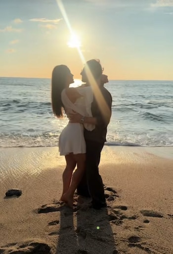 Una pareja se abraza en una playa de arena. El sol se pone sobre el océano, creando un resplandor brillante. Hay olas y huellas en la arena