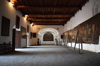 Interior de sala de museo con techo de madera, paredes blancas, cuadros de gran formato sobre caballetes y colgados, suelo de piedra, dos esculturas y una banca de madera