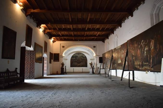 El Museo de Arte Colonial exhibe grandes pinturas en una sala de techos con vigas de madera y suelo de piedra, junto a esculturas y una banca de madera. (Agencia Guatemalteca de Noticias)