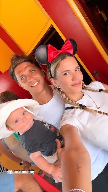 Un hombre y una mujer sonríen con un bebé en brazos. La mujer usa orejas de Minnie Mouse; el bebé, sombrero blanco y camiseta de Mickey Mouse. Fondo amarillo y rojo