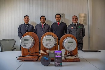Cuatro hombres posan detrás de una mesa con tres medidores de electricidad en soportes de madera, un dispositivo redondo y un smartphone con una aplicación