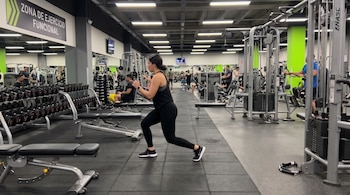 Una mujer con ropa deportiva negra hace desplantes en un gimnasio. Se observan equipos de ejercicio, mancuernas, bancos y otras personas entrenando.