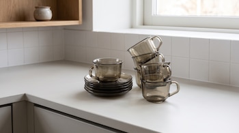 Varias tazas de vidrio ahumado y una pila de platos pequeños sobre una encimera clara de cocina frente a una pared de azulejos blancos.