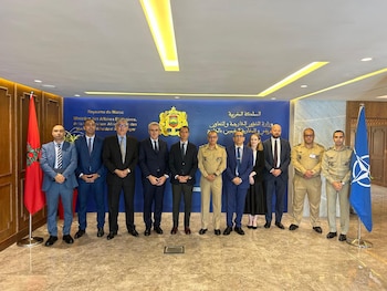 Un grupo de personas en traje civil y uniforme militar posa entre la bandera de Marruecos y la bandera de la OTAN ante un mural azul