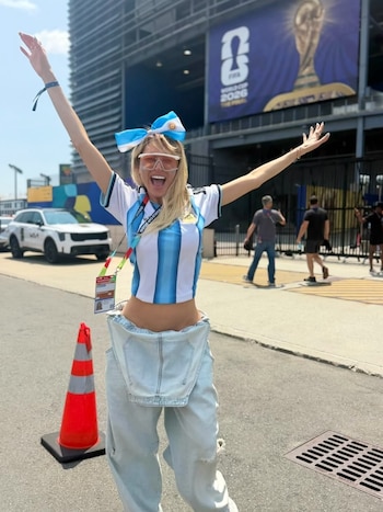 Una mujer con camiseta de Argentina, overalls y lazo celeste y blanco levanta los brazos frente a un estadio con el logo FIFA World Cup 2026 The Final