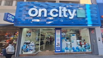 Fachada azul de tienda con letreros 'on city' en blanco, vitrinas con televisores, consolas, refrigeradores, globos celestes y blancos, personas