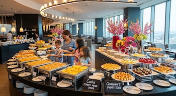 Mujer y niños se sirven en un buffet de restaurante moderno. Hay bandejas de waffles, postres, comida caliente y flores, con vista panorámica a la ciudad.