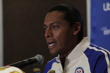 Primer plano de Joel Huiqui, un hombre con piel morena y cabello oscuro, hablando frente a un micrófono. Viste una chaqueta deportiva blanca y azul con franjas
