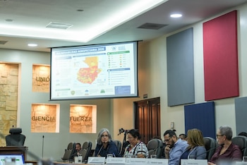 Un grupo de personas sentadas en una mesa de conferencias frente a una pantalla grande que muestra un mapa de Guatemala y datos sobre vulnerabilidad agropecuaria