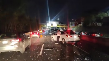 Carretera con tráfico de vehículos detenidos por la noche. Un coche blanco tiene la puerta del conductor abierta y un hombre está de pie. Restos en el asfalto.