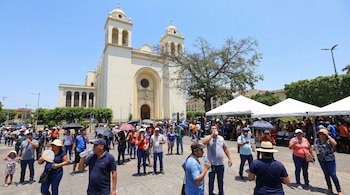 Una multitud de personas al aire libre bebiendo agua y usando sombrillas frente a la Catedral Metropolitana de San Salvador bajo un cielo azul claro.