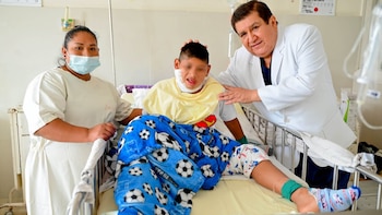 Un médico y una enfermera rodean a un niño en una cama de hospital. El niño usa vendajes en el cuello, una bata amarilla y una manta azul con balones