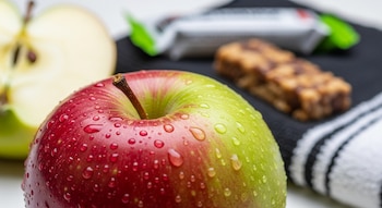 Una manzana entera roja y verde con gotas de agua, media manzana cortada, una barra de energía y una toalla de rayas.