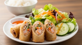 Un plato blanco muestra rollitos de primavera, algunos abiertos con camarones y vegetales, salsa, ensalada de hojas verdes, zanahoria y pepino. Arroz blanco al fondo.