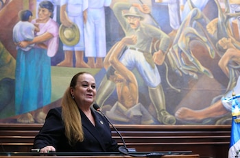 Una mujer con cabello castaño claro habla en un podio con micrófono. Detrás, un mural colorido muestra figuras humanas en distintas poses, incluyendo algunas en conflicto