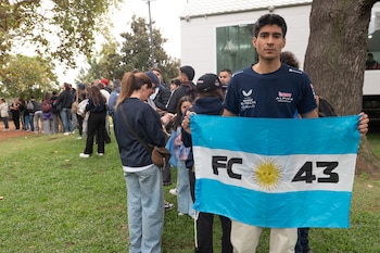 Un hombre joven con camiseta oscura y pantalón claro sostiene una bandera argentina celeste y blanca con las letras "FC" y el número "43". Al fondo, hay personas y árboles