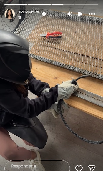 Primer plano de María Becerra, con casco de soldar negro y guantes grises, soldando un perfil metálico unido a una reja de malla, con unas tenazas rojas cerca