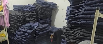 Una habitación con numerosas pilas altas de prendas de vestir oscuras plegadas, posiblemente chaquetas, apiladas contra paredes claras y en el suelo