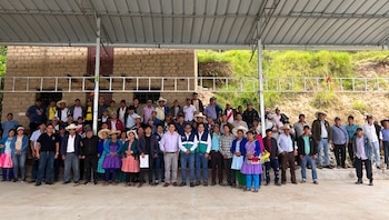 Un gran grupo de personas, hombres y mujeres de diversas edades, posa para una foto bajo un techo metálico, frente a un edificio de adobe y un paisaje verde
