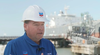 Andy Walz, ejecutivo de Chevron, vestido con casco blanco y uniforme azul, habla frente a un puerto industrial con un gran barco borroso