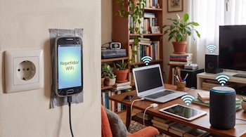Teléfono adherido a una pared con la frase "Repetidor WiFi" en pantalla, cerca de una mesa con computadora, tableta y parlante inteligente.
