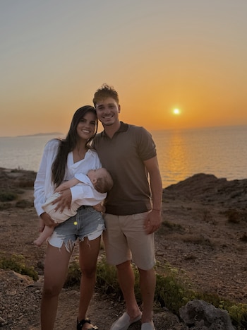 Un hombre, una mujer y un bebé posan en un terreno rocoso. La mujer sostiene al bebé. Al fondo, el mar y una puesta de sol naranja