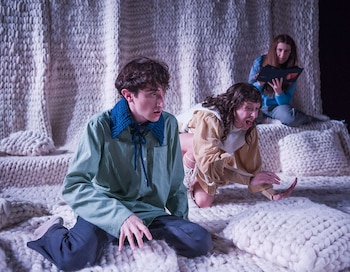 Tres actores en una escenografía de tejido blanco. Un joven arrodillado, una mujer agachada con la boca abierta y otra mujer sentada al fondo leyendo un libro