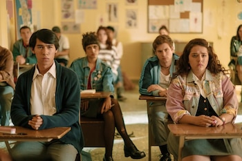 Jóvenes sentados en pupitres de madera en un aula. Hay tres estudiantes en primer plano mirando al frente. Más personas y afiches en la pared al fondo