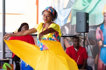 Una mujer afroamericana sonriente baila con un vestido amarillo brillante, un top floral y un pañuelo en la cabeza, frente a músicos y un mural colorido