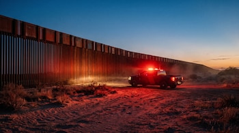 Camioneta con luces rojas encendidas en un camino de arena al lado de una pared metálica alta durante el crepúsculo en el desierto.