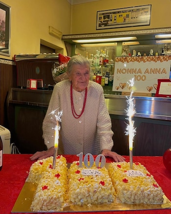 Mujer mayor con collar rojo detrás de una torta de cumpleaños con el número cien, las palabras 'AUGURI ANNA' y dos bengalas. Detrás, un cartel 'NONNA ANNA 100'