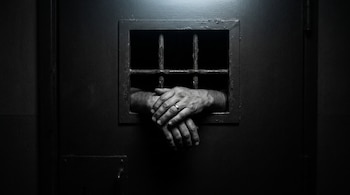 Dos manos de hombre con un anillo, entrelazadas, que asoman por una ventana enrejada de una puerta de celda, rodeadas de oscuridad.