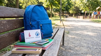 Mochila azul, útiles escolares, cuadernos, lápices, calculadora, estuche, tarjeta con logos ICHMUJERES y CEAVE, banco de madera en parque.