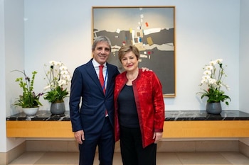 Luis Caputo, con traje oscuro y corbata roja, junto a Kristalina Georgieva, con chaqueta roja, posando frente a una pintura abstracta y plantas de orquídeas