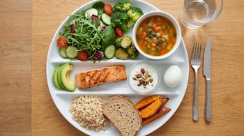 Vista aérea de un plato dividido con ensalada, brócoli, coles de Bruselas, sopa de verduras, salmón, aguacate, yogur con frutos secos, huevo, arroz integral, pan y batatas.