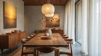 Una habitación con una mesa de comedor de madera larga rodeada de sillas de madera, dos lámparas colgantes, cuadros de paisajes y cortinas blancas.