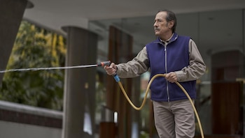 Guillermo Francella, un hombre de mediana edad con bigote, camisa beige y chaleco azul, rocía agua con una manguera amarilla y boquilla roja