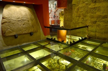 Interior de museo con piso de vidrio, vitrinas con rocas y artefactos, estela de piedra tallada en pared y escultura dorada sobre pedestal