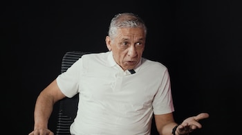 Hombre mayor de cabello gris con camisa polo blanca sentado en silla de rayas grises, fondo negro liso, brazo derecho extendido y micrófono en solapa.